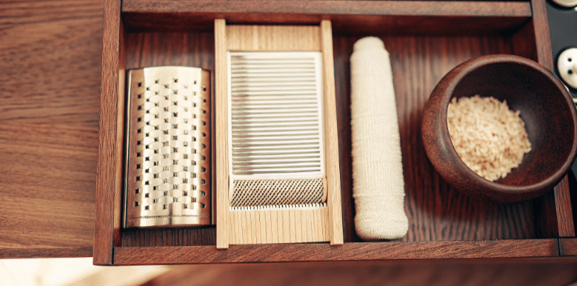 The image showcases essential Japanese kitchen tools for grating and grinding, including a traditional Japanese grater for ginger and daikon radish, a sesame seed grinder for creating rich paste, and a mortar and pestle for crushing spices. These utensils are vital for enhancing flavors in authentic Japanese dishes like miso soup and omelets.