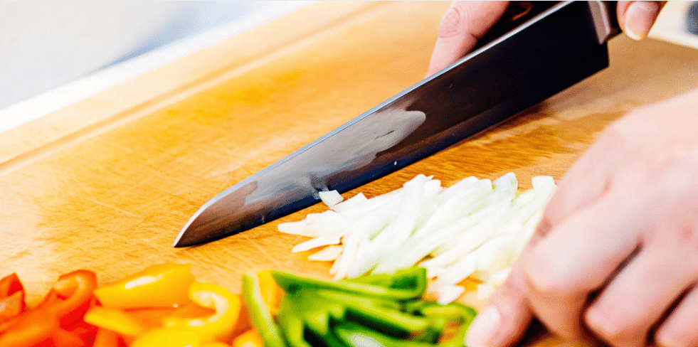 A person is demonstrating the proper technique for using a nakiri knife, a Japanese vegetable knife, by holding it with a pinch grip and performing an up and down chopping motion on a cutting board. The image showcases the knife's straight blade and rectangular shape, emphasizing its effectiveness for cleanly slicing vegetables like onions and bell peppers.