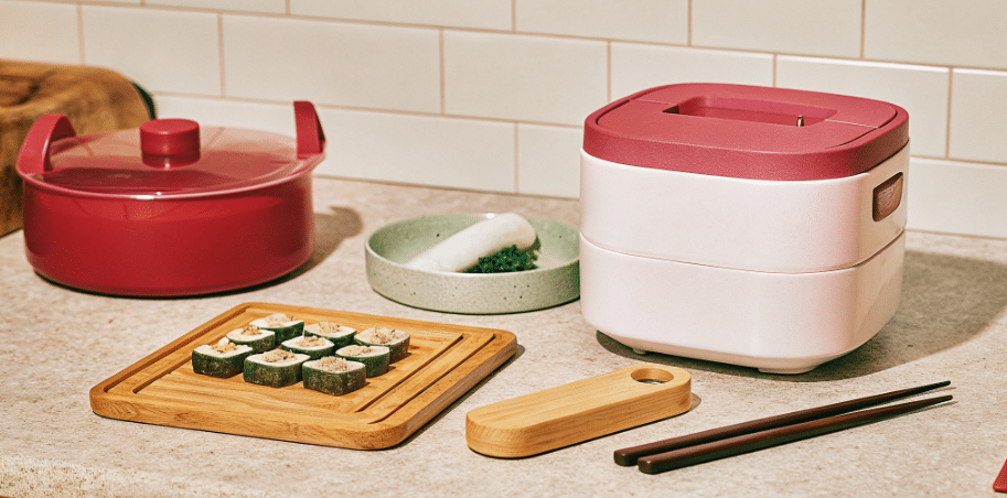 The image depicts a well-organized Japanese kitchen featuring essential Japanese cooking tools, including a rice cooker, a sushi oke for mixing sushi rice, and a Japanese-style mortar and pestle for grinding spices. Long chopsticks, a drop lid for simmering soups, and a soft wooden cutting board are also visible, highlighting the unique equipment needed for preparing authentic Japanese dishes like sushi rolls and miso soup.