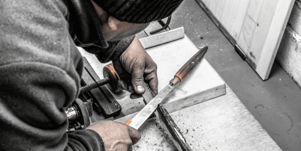 Making Your Own Kogatana - Japanes Knives Tips The image depicts a craftsman working on a kiridashi knife, showcasing the process of shaping high carbon steel into a blade with a precise cutting edge. Tools such as a grinder, file, and heat source are visible, emphasizing the detailed craftsmanship involved in creating this versatile tool.