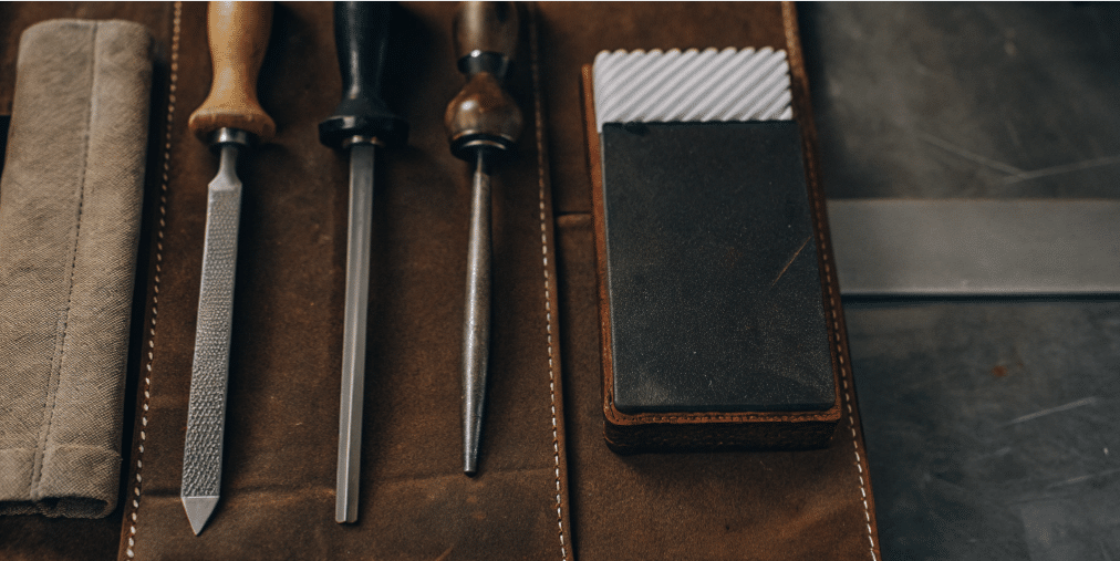 The image shows various tools for sharpening serrated knives, including a tapered diamond rod and honing steel, which are designed to fit into the scallops of serrated edges. Additionally, there is a leather strop and finer grit stone for refining and polishing the edge after sharpening, ensuring the knives maintain their sharpness and cutting ability.