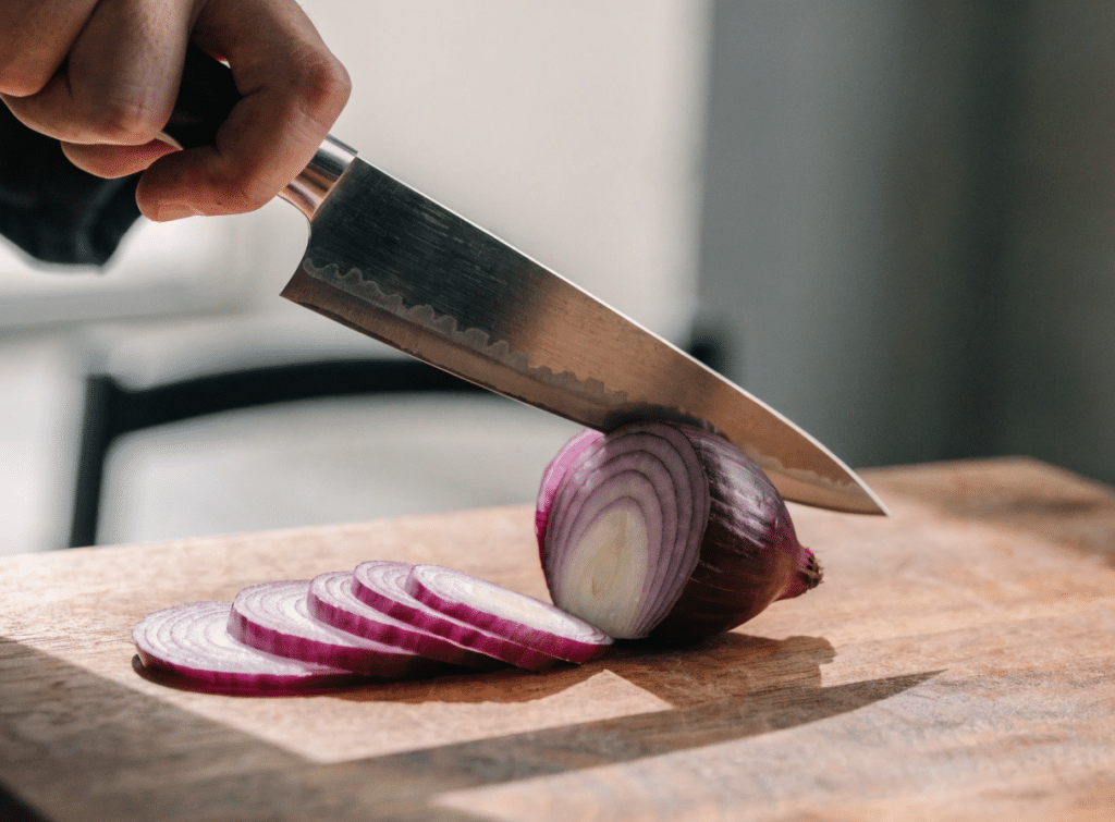 Japanese Knife Cutting Onion - Japanes Knives Tips A close-up photo of a Japanese chef knife slicing a red onion into very thin even slices on a wooden board, sharp edge detail, clean kitchen setting, natural light, shallow depth of field, editorial food photography style